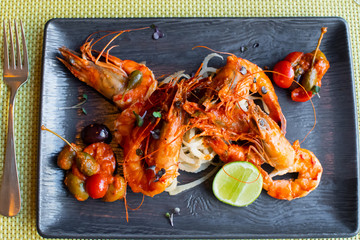 Sauteed shrimps in tomato, capers and olives sauce. Black plate in a restaurant setting background