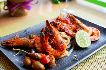 Sauteed shrimps in tomato, capers and olives sauce. Black plate in a restaurant setting background