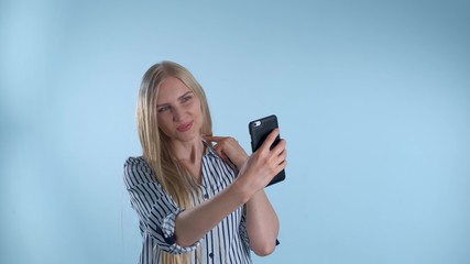 Attractive blonde lady making selfie on smartphone on blue background. She posing and smiling to the camera.