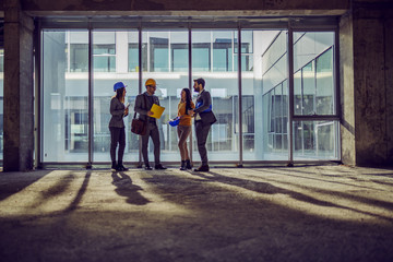 Four dedicated hardworking architects standing next to big window in and old building and talking about how they plan to rebuilt it to be modern. - obrazy, fototapety, plakaty