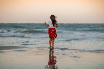 Fototapeta na wymiar little girl in a red skirt in white peas runs along the seashore