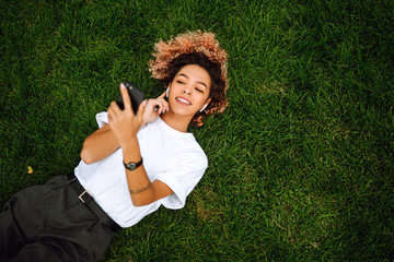 Smiling woman using a smart phone and relaxing at the park on green grass. Concept of leisure,...