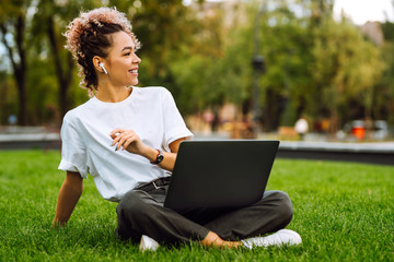 Woman  wearing earphones sitting on grass at park working on laptop. Freelance, blogging or remote work concept.