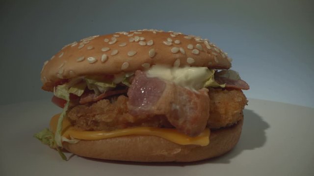 A delicious appetizing cheeseburger with meat, sausage, tomato, cheese and greens rotates on a white surface. Loop