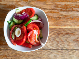 top view of homemade salad from sliced fresh vegetables (oiled tomato, cucumber, onion) in white bowl on shabby wooden table at home kitchen