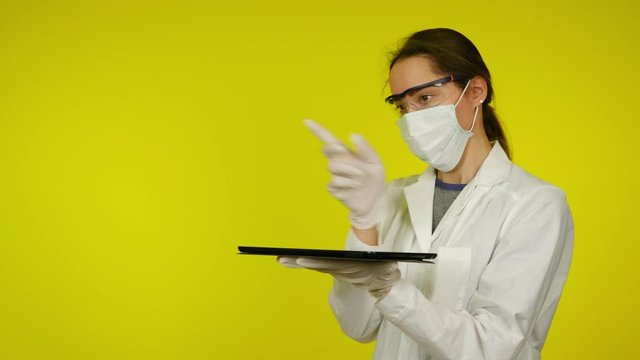 Woman in medical coat and protective mask uses holographic technology of the future on tablet screen. Augmented reality concept. Yellow background with copy space. Place for graphics. 4K footage