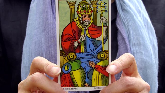 A woman holding "The Pope", Tarot de Marseille, walks to the camera in front of waterfall, Tokyo, Japan. April 2020. Camera fixed, close-up, focus in, neutral level.