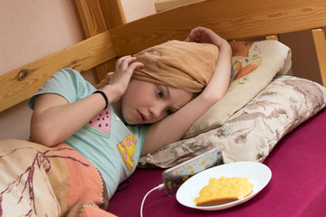 A teenager girl after a shower or taking a bath lies in bed under a blanket with her hair wrapped in a towel. A girl looks at the phone and eats a snack cake in bed.