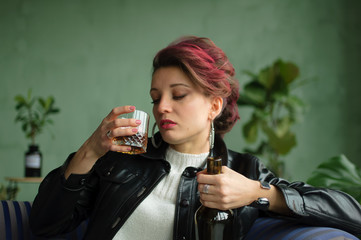 Young sad woman with dark pink hair with glam rock style look is drinking whisky from a glass on...