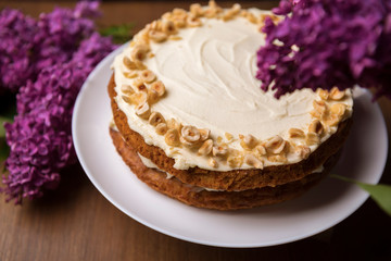 Carrot cake with hazel nuts and bouquet of purple lilac on the wooden table