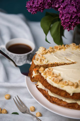 Carrot cake with hazel nuts, bouquet of purple lilac and cup of coffee on the white tablecloth