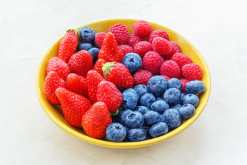 Strawberries raspberries blueberries in a yellow bowl on a light background, selective focus