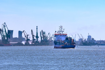 View of commercial cargo port with container bridge cranes and a ship entering port on a sunny day.