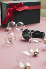 Black wristwatch on a pink color background, near gift boxes tied with a red ribbon. Glass balls.