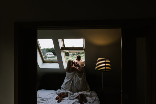 Young man and woman in sheets hug and kiss near the window. Photo nude men and women at the window.