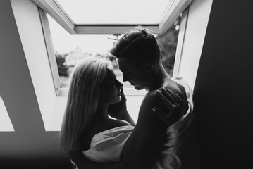 Young man and woman in sheets hug and kiss near the window. Black and white photo of a naked man and woman by the window.