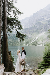 Beautiful bride in a boho style dress and groom are hugging near the lake against the backdrop of the mountains. Wedding photo shoot in the mountains. Black and white photo of a bride and groom in the