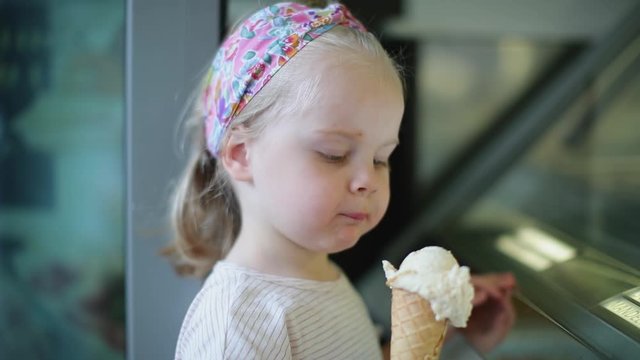 Little blonde stands near the window and eats ice cream 