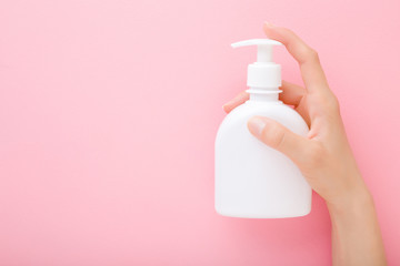 Young woman hand holding white pump bottle on light pink table background. Pastel color. Care about clean and soft body skin. Daily beauty product. Closeup. Empty place for text or logo. Top view.