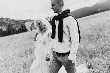 Beautiful bride in a boho style dress and groom walk on the field near the mountains. Wedding photo shoot in the mountains. Black and white photo of a bride and groom in the mountains.