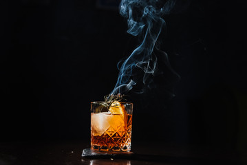 Whiskey with orange, ice and smoking rosemary close-up.