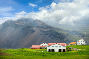 Many white buildings and red roofs are factories and pastures. In the countryside of Iceland in the summer there will be green grass. The background is large and tall mountains and blue sky.