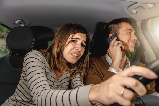 Young Man Talking On The Phone While Driving And Woman Passenger Grabs Steering Wheel Trying To Avoid An Obstacle. Scared And Screaming Couple Is Getting A Car Accident.