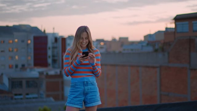 Cute young woman in colourful clothes on warm summer evening or dawn laughs and smiles, look at phone.City skyline in background in soft paster light.Young zoomer generation stay connected in internet