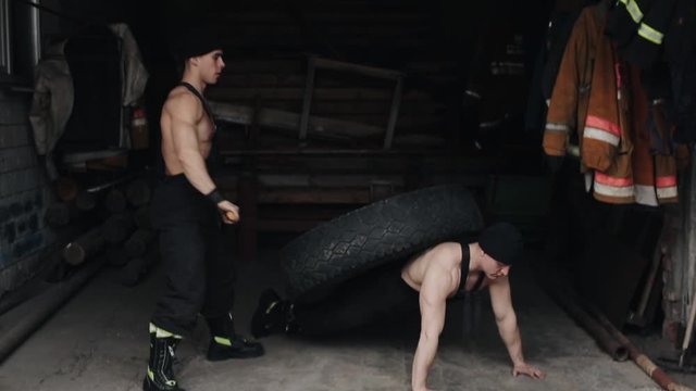 Two muscular young men are training and doing extreme crossfit exercises with a tire and a sledgehammer. The camera moves up and down