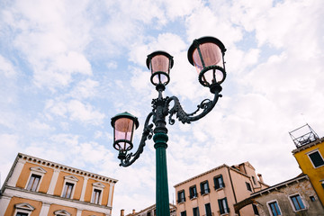 Fototapeta na wymiar An ancient triple metal street lamp, with pink plafonds against the sky and buildings of Venice, Italy. Vintage street lighting, elegant pillar design.