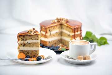 Cup of coffee with two pieces of sugar on a saucer and next to a carrot cake covered with caramel and decorated with nuts on top, behind a white textile background