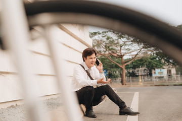 Fashionable young bearded man using a digital tablet , cheerfully smiling, watching or reading something funny, sitting on the ground, near the bicycle, outdoors.