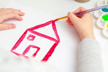 Child is drawing house roof with watercolors on white paper.