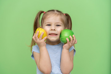 Fototapeta na wymiar little girl 4 years old in a blue T-shirt on a green background put an apple and a lemon to her face and smiles. pre-school children healthy eating concept