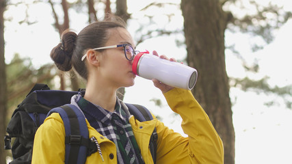 Young girl tourist in yellow jacket drinking tea from a thermos. Female hiker with backpack enjoying nature
