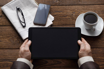 Top view on male hands holding a tablet, a cup of coffee and a morning newspaper. Work is all...