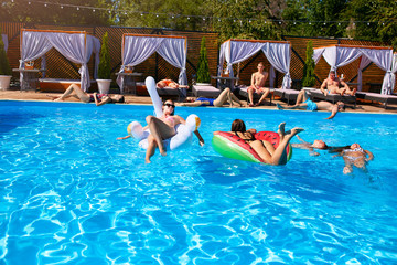 Group of friends chilling in private villa swimming pool lie in the sun on inflatable flamingo, swan, mattress. Young people relax with floaties at luxury resort on sunny day. Bikini girls sunbathing.