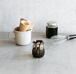 Homemade dark chocolate sauce in glass jars, ice cream waffle cones and silver whisk on gray background. 