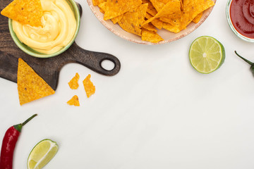 top view of corn nachos with chili pepper, lime, ketchup, cheese sauce on wooden cutting board on white background