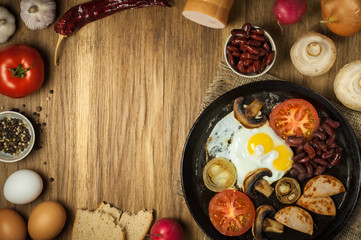 fried eggs with ham, onion, tomatoes, mushrooms and red beans in a pan on an old wooden surface together with raw ingredients. Creative rustic layout with a set of products and copy space. top view