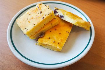 four square pieces of yellow cheesecake sprinkled with sesame seeds and flax on a white and blue-rimmed round plate on a wooden table . gluten-free homemade cakes