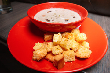 Beige cream vegetable and mushroom soup in a red  bowl and plate on a black table.