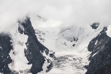 Atmospheric minimalist textured alpine landscape with massive glacier on big mountain in low clouds. Background of snowbound mountainside. Cracks on ice. Majestic foggy misty scenery on high altitude.