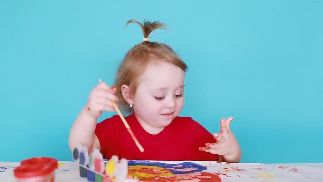 A cheerful little blonde girl draws with a brush and colored paints on a white canvas. The child with the ponytail was covered in paint. Space for text