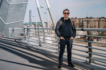 A young man in a jacket and glasses stands on a bridge, beautiful metal structures in the air, hair flying in the wind, business issues, construction engineering