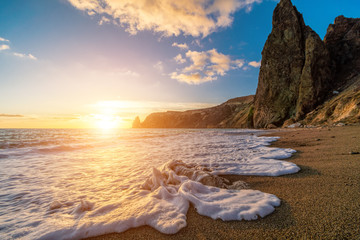 Bright sea sunset. The waves crash into the rock, lit by the warm sunset, sand and pebbles,...