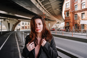 Portrait of a girl with long hair in a coat and a backpack standing on a street in St. Petersburg, a brick old building and bridge beams located close to a residential building
