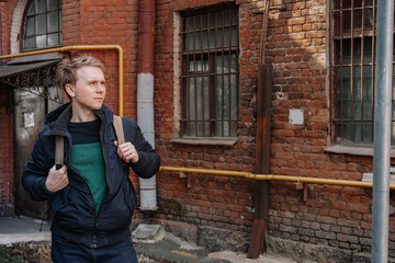 Portrait of a yoyng man with blond hair in a coat and a backpack standing on a street in St. Petersburg, a brick old building and bridge beams located close to a residential building