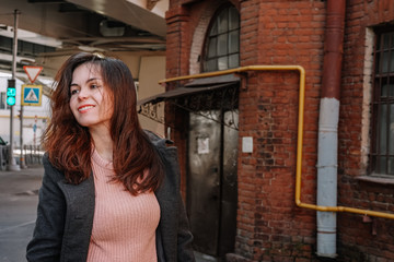 Portrait of a girl with long hair in a coat and a backpack standing on a street in St. Petersburg, a brick old building and bridge beams located close to a residential building