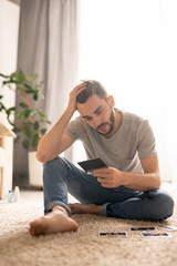 Lonely young bearded man in jeans sitting on carpet and holding head in hand while watching old photos in home isolation